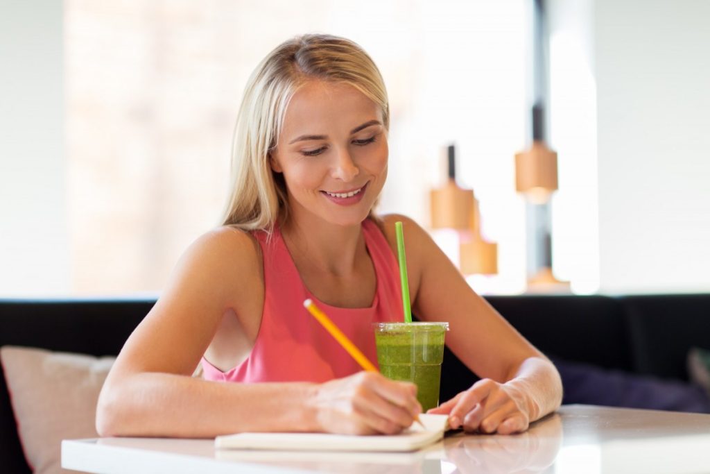 13 Ways To Look 10 Years Younger Naturally Without Costing The Earth; woman with drink writing to notebook at restaurant